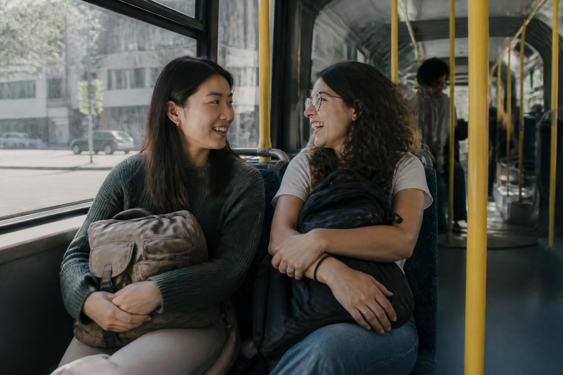 Two students laughing together on a city bus with their backpacks